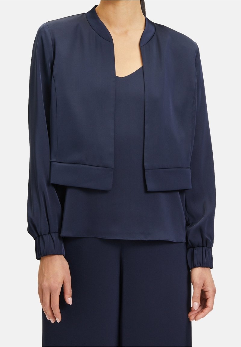 Femme portant une veste courte bleu marine à manches longues sur un top sans manches assorti et un pantalon à jambes larges, avec des poignets froncés sur la veste.