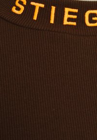 Brązowa prążkowana tkanina z pomarańczowym haftowanym napisem wzdłuż kołnierza, ukazująca zbliżenie detali tekstury i szwów.