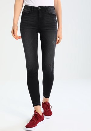 Personne portant un jean skinny noir légèrement usé et des baskets rouges, debout devant un fond clair uni.