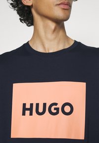 Námořnické modré bavlněné tričko s velkým broskvovým obdélníkovým grafickým prvkem a textem "HUGO" v tučných černých písmenách. Klasický design s kulatým výstřihem.