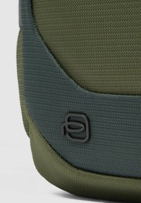 Materiale tessile verde con superficie texturizzata. Include un accento logo nero e un bordo in tessuto verde a contrasto.