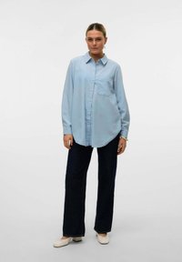 Svijetloplena košulja s dugim rukavima, šiljastim ovratnikom, prednjim džepom i opuštenim krojem, uparena s tamnoplavim širokim trapericama.