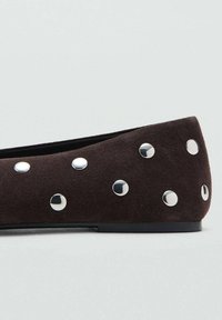 Zapato plano de ante marrón oscuro con tachuelas redondas de plata distribuidas uniformemente a lo largo del lateral y la parte trasera, suela negra y tacón bajo.