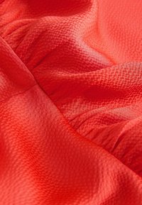 Tkanina z teksturo v živahni rdeči barvi, z gladkimi in nabrani deli, ki ustvarjajo globino in vizualni učinek. Lahka in tekoča zasnova.