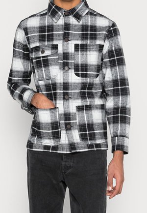 Man draagt een zwart-wit geruit overhemd met knopen en borstzakken, samen met een donkere broek, hand in de zak.