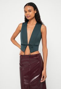 Frau mit langen lockigen Haaren trägt eine tiefgrüne, ärmellose Weste, die an der Taille gebunden ist, und einen glänzenden dunkelroten Lederrock.