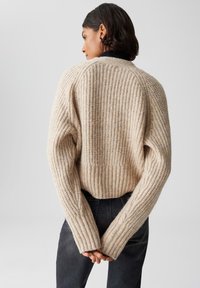 Donna con capelli corti scuri che indossa un maglione beige a maglia grossa e jeans neri, in piedi con le mani giunte dietro la schiena su uno sfondo grigio.