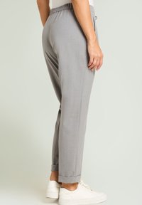 Graue maßgeschneiderte Hose mit Kordelzug in der Taille, zulaufendem Bein und umgeschlagenem Saum. Kombiniert mit weißen Schuhen vor hellem Hintergrund.