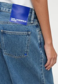 Τζιν σε ανοιχτό μπλε χρώμα, με ορθογώνια μπλε δερμάτινη ετικέτα στο πίσω μέρος, στην οποία είναι αποτυπωμένο το "KARL LAGERFELD JEANS" σε λευκό.