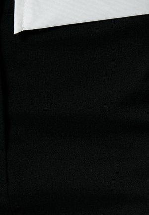 Tessuto nero dei pantaloni con una superficie liscia, con una camicia bianca sottostante, parzialmente visibile lungo il bordo superiore.