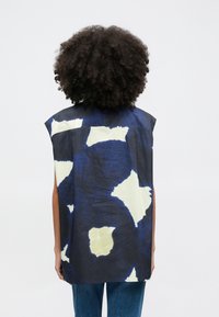 Ujjatlan top sötétkék anyagból, szabálytalan krémszínű mintákkal; egyenes vágású, látható oldalzsebekkel a kényelmesebb illeszkedésért.