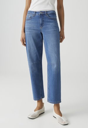 Donna con jeans blu a gamba dritta e scarpe bianche intrecciate slip-on, in piedi davanti a uno sfondo grigio uniforme.