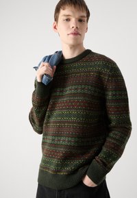 Green and brown patterned sweater with multicolored accents. Ribbed neckline and cuffs. Model holds denim fabric.