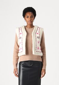 Cream faux-fur vest with pink and black embroidered stars, layered over a ribbed beige sweater. Model wears a black leather skirt.