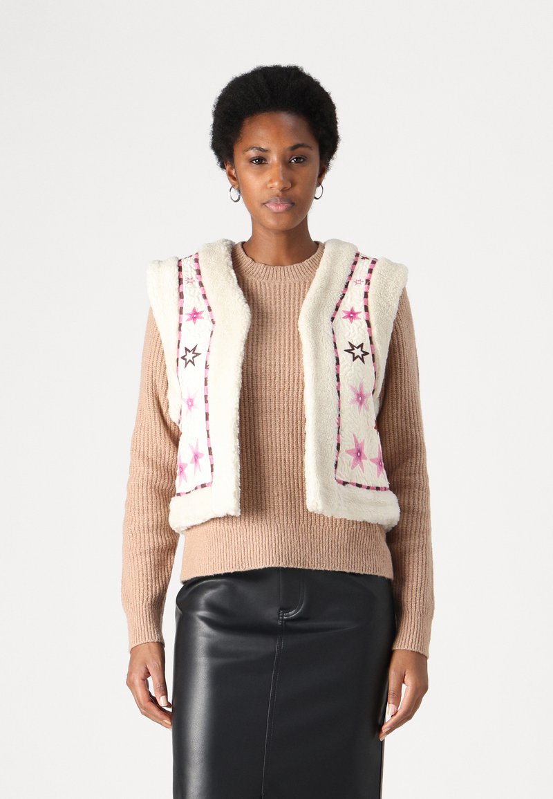 Cream faux-fur vest with pink and black embroidered stars, layered over a ribbed beige sweater. Model wears a black leather skirt.