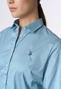 Camicia azzurra chiaro con colletto, realizzata in tessuto liscio; presenta chiusure con bottoni e un piccolo logo navy ricamato nella zona sinistra del petto.