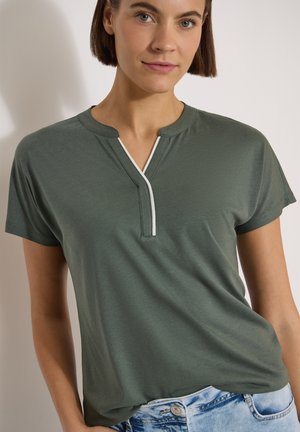 Femme aux cheveux courts et bruns portant un t-shirt vert à manches courtes avec un col en V bordé de blanc et un jean bleu clair.