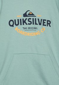 Light mint green pullover sweatshirt with a front pocket. Features "QUIKSILVER" logo in black, accented with orange text. Soft fabric, casual design.