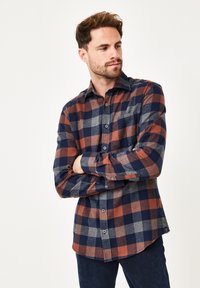 Mann mit kurzen braunen Haaren und Bart, der ein kariertes Button-Up-Shirt in Orange, Navy und Grau trägt, mit verschränkten Armen, nach unten rechts auf einem weißen Hintergrund blickend.