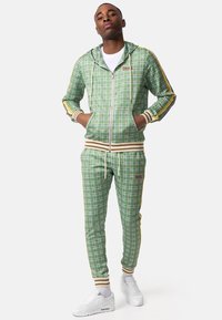 Green plaid zip-up hoodie with beige and yellow stripes on sleeves, matching fitted pants, and white sneakers. Fabric has a smooth texture.