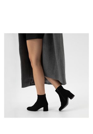 Jambes portant des bottines noires à talons, une jupe noire et un long manteau gris marchant sur une surface blanche.