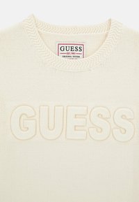 Crème kleurige gebreide trui met een textuur, verhoogd "GUESS" logo aan de voorkant en een eenvoudige ronde halslijn met ribbel detail.
