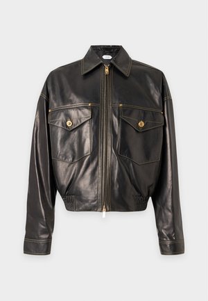 Junger Mann trägt eine schwarze Lederjacke, braunes Hemd, gestreifte blaue und braune Hose und schwarze Schuhe mit goldenen Details, stehend vor einem einfarbigen Hintergrund.