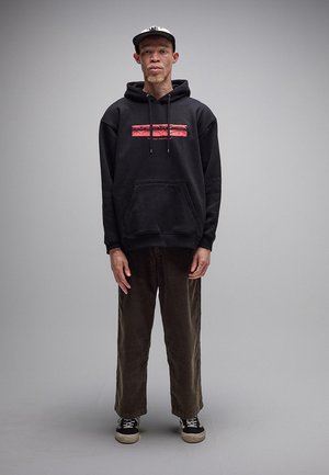 Junger Mann steht, trägt schwarzen Hoodie mit rotem Grafikprint, dunkle Hose, schwarze Turnschuhe und schwarze-weiße Kappe vor einfarbig grauem Hintergrund.