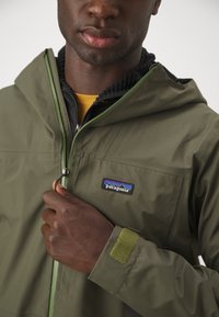 Veste imperméable olive avec capuche, fermeture éclair verte contrastante et logo Patagonia. Dispose d'un poignet en velcro et d'un doublure intérieure texturée.