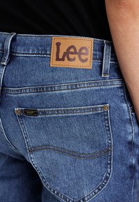 Traperice srednje plave boje s teksturiranim završetkom. Istaknuti tamni šavovi i smeškan kožni oznaka sa "Lee" logotipom na pojasu.