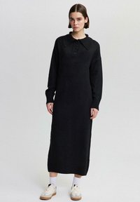 Abito nero lavorato a maglia con un ampio colletto, maniche lunghe e chiusura frontale con bottoni, caratterizzato da polsi e orlo a coste. Indossato con sneaker beige.