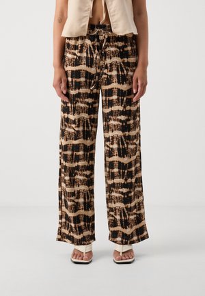 Mujer que lleva pantalones anchos beige con estampado negro, top corto beige y sandalias blancas de punta cuadrada, de pie frente a un fondo blanco.