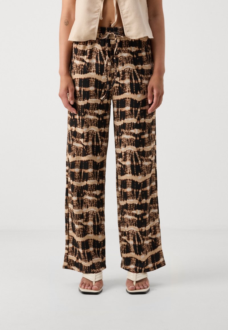 Femme portant un pantalon large à motifs beige et noir, un top court beige et des sandales blanches à bout carré, debout devant un fond blanc.