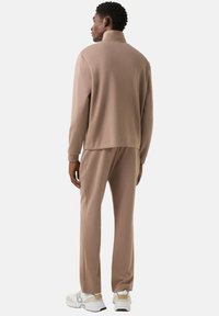 Světlě hnědá tepláková souprava z měkké látky; má vysoký límec, dlouhé rukávy a zúžené kalhoty. Nošená s bílými teniskami.