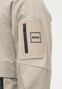 Μανίκι από μπεζ σακάκι με ορθογώνιο μπεζ τμήμα που διαθέτει μαύρο κατακόρυφο φερμουάρ και λογότυπο "BOSS" μέσα σε μαύρο πλαίσιο.