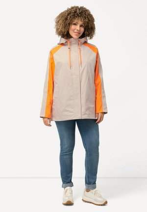Kvinde med krøllet hår iført en lysebeige jakke med orange striber, blå opkørte jeans og beige sneakers, stående mod en hvid baggrund.