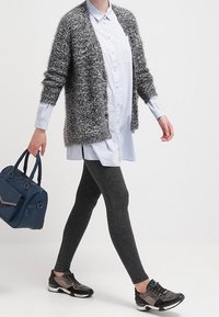 Grå fluffig cardigan över en ljusblå skjorta med knappar, tillsammans med svarta texturerade leggings och mörka sportskor. Blå handväska i handen.