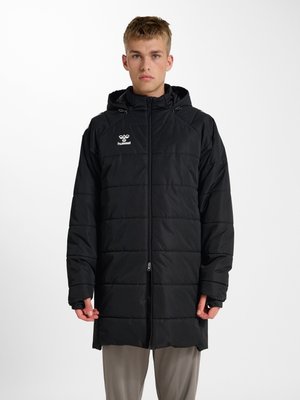 Jonge persoon die een lange zwarte gewatteerde jas met capuchon draagt, staand tegen een effen witte achtergrond.