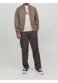 Chaqueta bomber de ante marrón con puños acanalados, combinada con una camiseta gráfica crema y pantalones gris oscuro; zapatillas en blanco y negro completan el look.