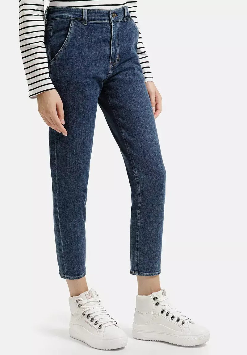 Niebieskie dżinsowe spodnie z wysokim stanem, zwężające się nogawki, przednie kieszenie oraz zamek błyskawiczny, zestawione z białymi trampkami za kostkę. Pasiasty top z długim rękawem.