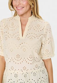 Cremefarbener Bluse mit Lochmustern, einer Rüschensilhouette am hochgeschlossenen Ausschnitt und Puffärmeln. Entworfen mit einem lockeren Schnitt und dezenten Mustern.