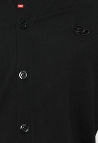 Melna adīta kardigāna ar trim melniem pogām, nelielu sarkanu etiķeti pie apkakles un melnu izšūtu olveida logotipu uz krūtīm.