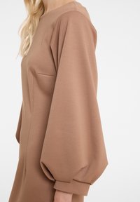 Femme portant une robe couleur camel avec de longues manches bouffantes et ajustées, vue de l'épaule jusqu'au milieu du bras.