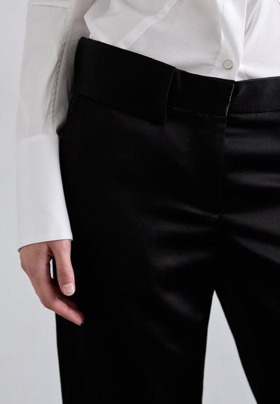 Pantaloni neri su misura con una texture liscia, dotati di una cintura alta e una chiusura frontale. Accompagnati da una camicia bianca con bottoni.