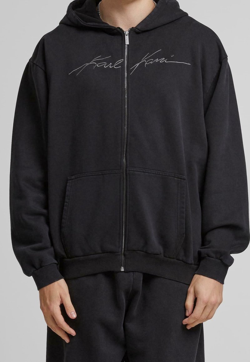 Personne portant un sweat à capuche noir zippé et un pantalon noir assorti, le sweat présente un logo cursif « Karl Kani » sur la poitrine.