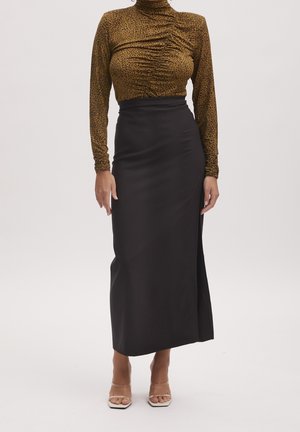 Vrouw met een bruin coltruitje met luipaardprint en een zwarte hoge taille midi-rok met een split, staand op een heldere achtergrond.