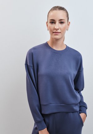 Jonge vrouw met een lichte huid en haar naar achteren gebonden, draagt een blauwe trui met lange mouwen en bijpassende broek, met één hand in haar zak.