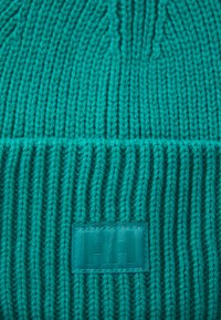 Knitted beanie in vibrant turquoise, featuring ribbed texture and a rectangular fabric tag with a logo.