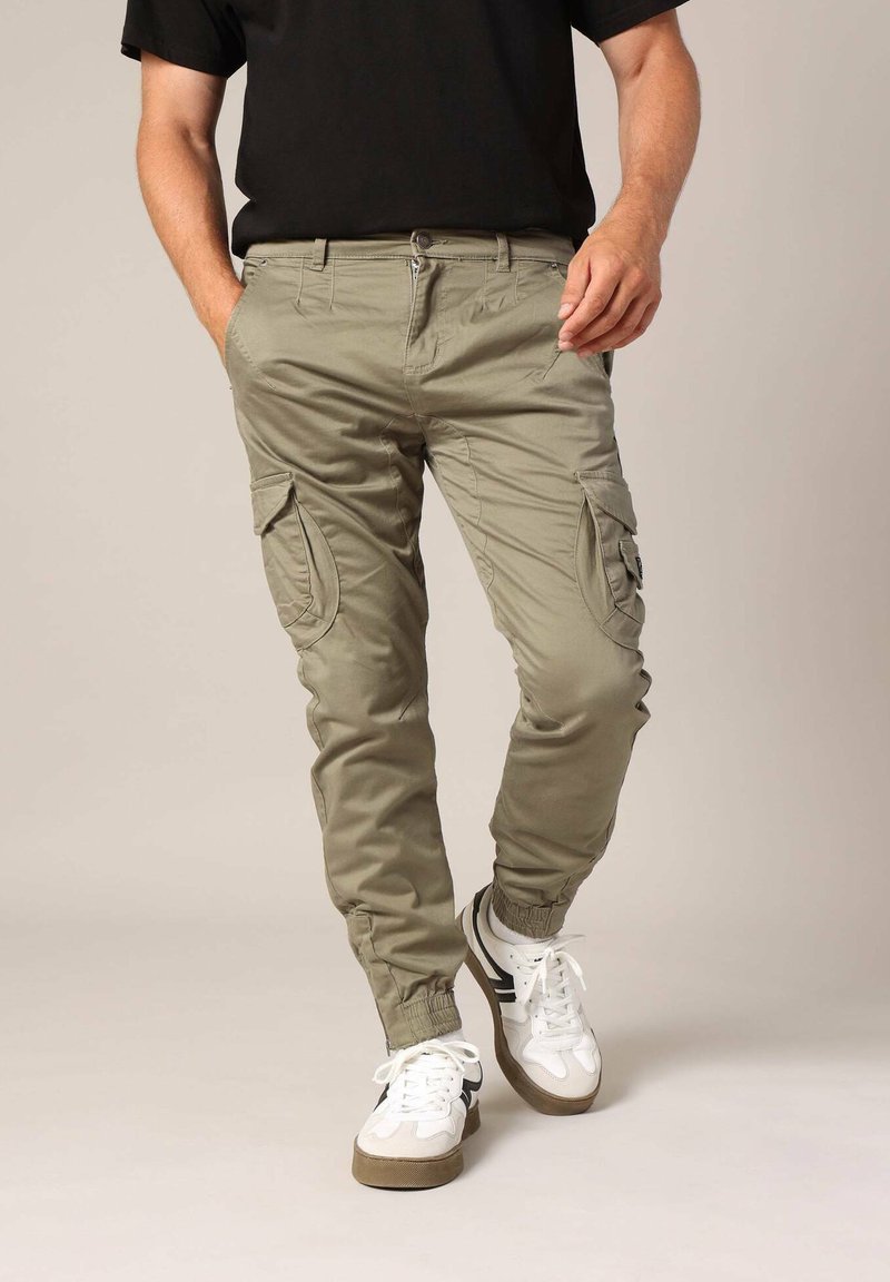 Khaki cargo pants met zijzakken, elastische boorden en een ritssluiting; gecombineerd met witte sneakers met zwarte accenten.