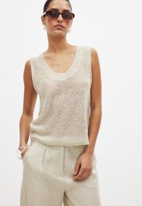 Light beige sleeveless knit sweater with a wide ribbed neckline, layered texture, and raw edge hem, paired with light beige linen trousers.